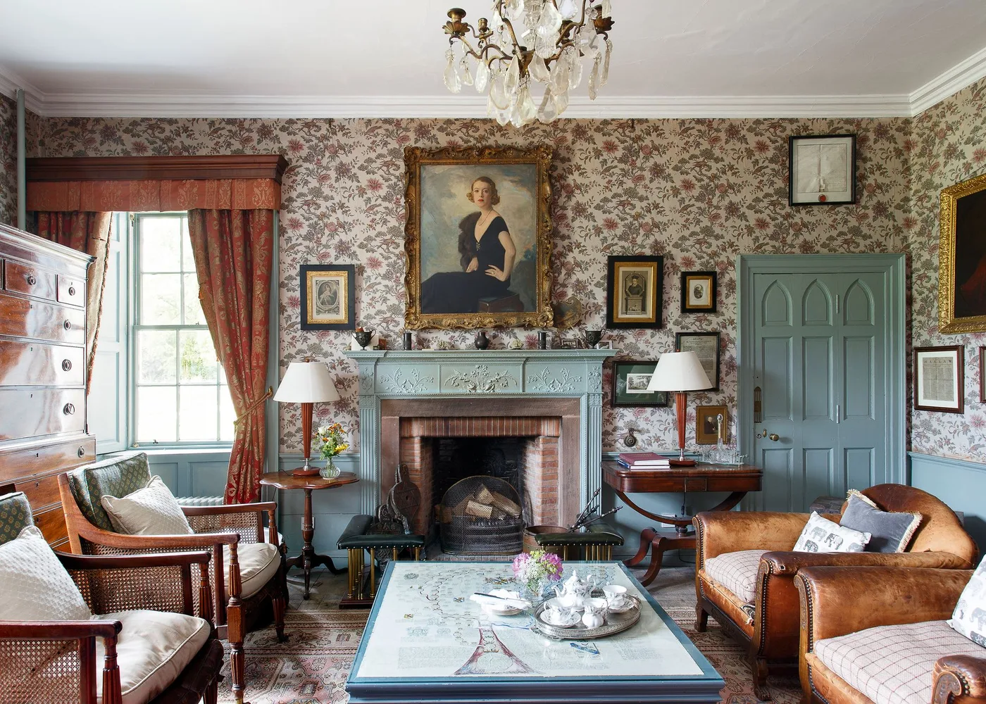 Floral-wallpapered sitting room with green carved mantel, central portrait, lamps and warm wood chairs