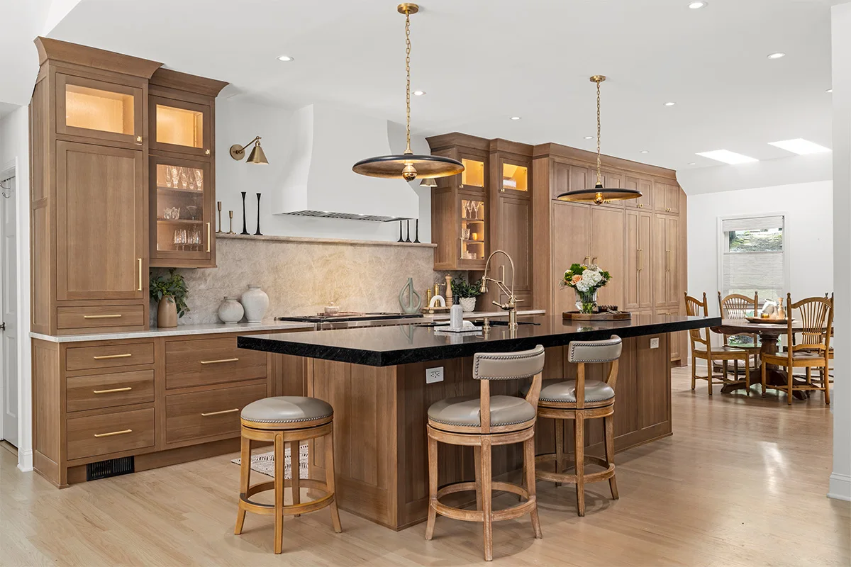 Brown stained oak cabinets with illuminated glass sections surround a pale hood, with a black island counter and round upholstered stools.