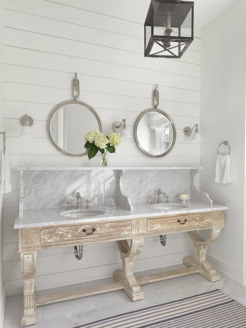 Pale bathroom with carved wood console double vanity, veined counter and backsplash, round mirrors, horizontal paneling and striped rug