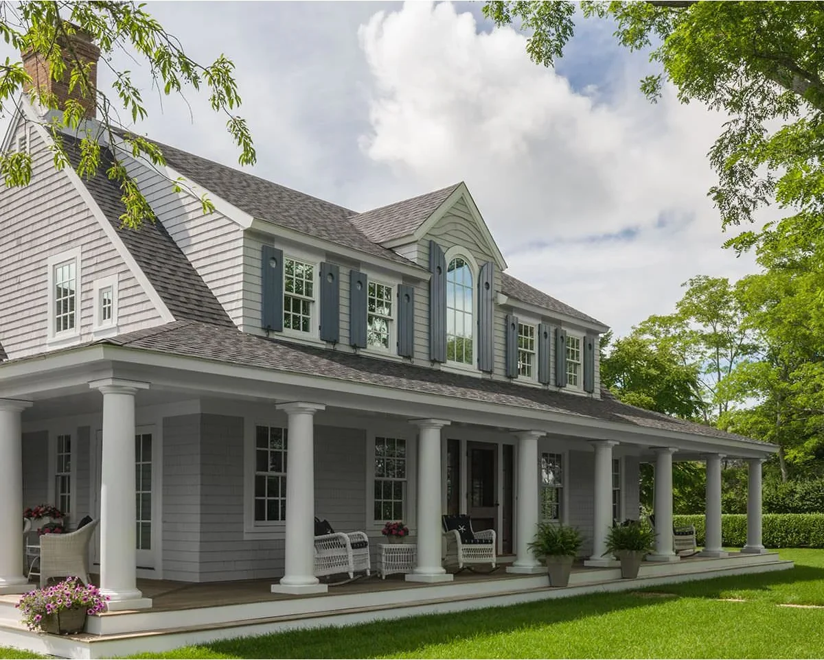 A large Cape-inspired shingle house has a broad wraparound porch with white columns, pale seating and darker upper-window shutters.