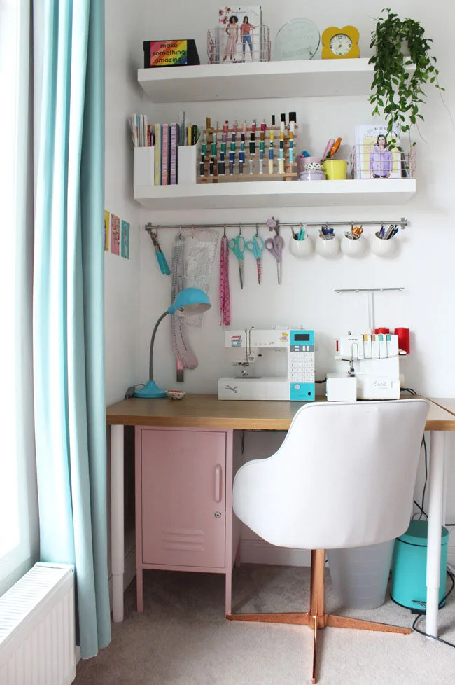 A sewing machine and overlocker sit on a wood worktop below tool rails and shelves, with a pale chair and pink cabinet underneath.