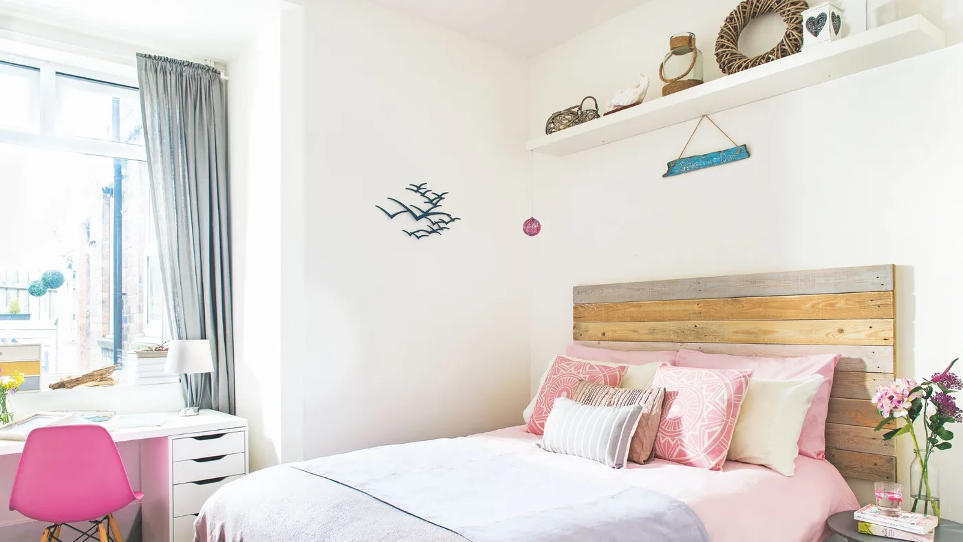 White bedroom with a horizontal wooden headboard, pale pink bedding, small nautical wall ornaments and a window-side desk.