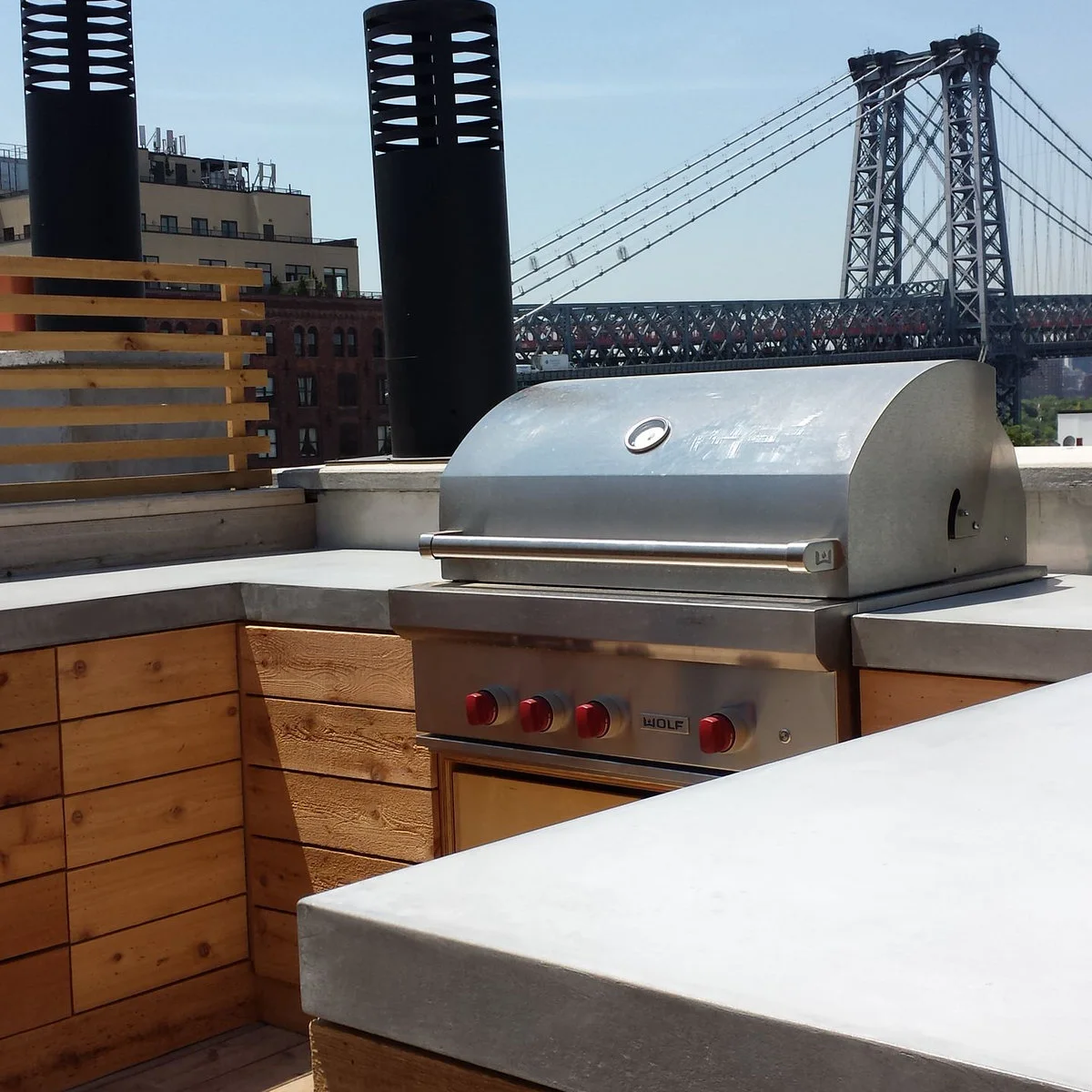 Rooftop outdoor grill area with horizontal wood cabinet fronts, pale counters and bridge visible beyond