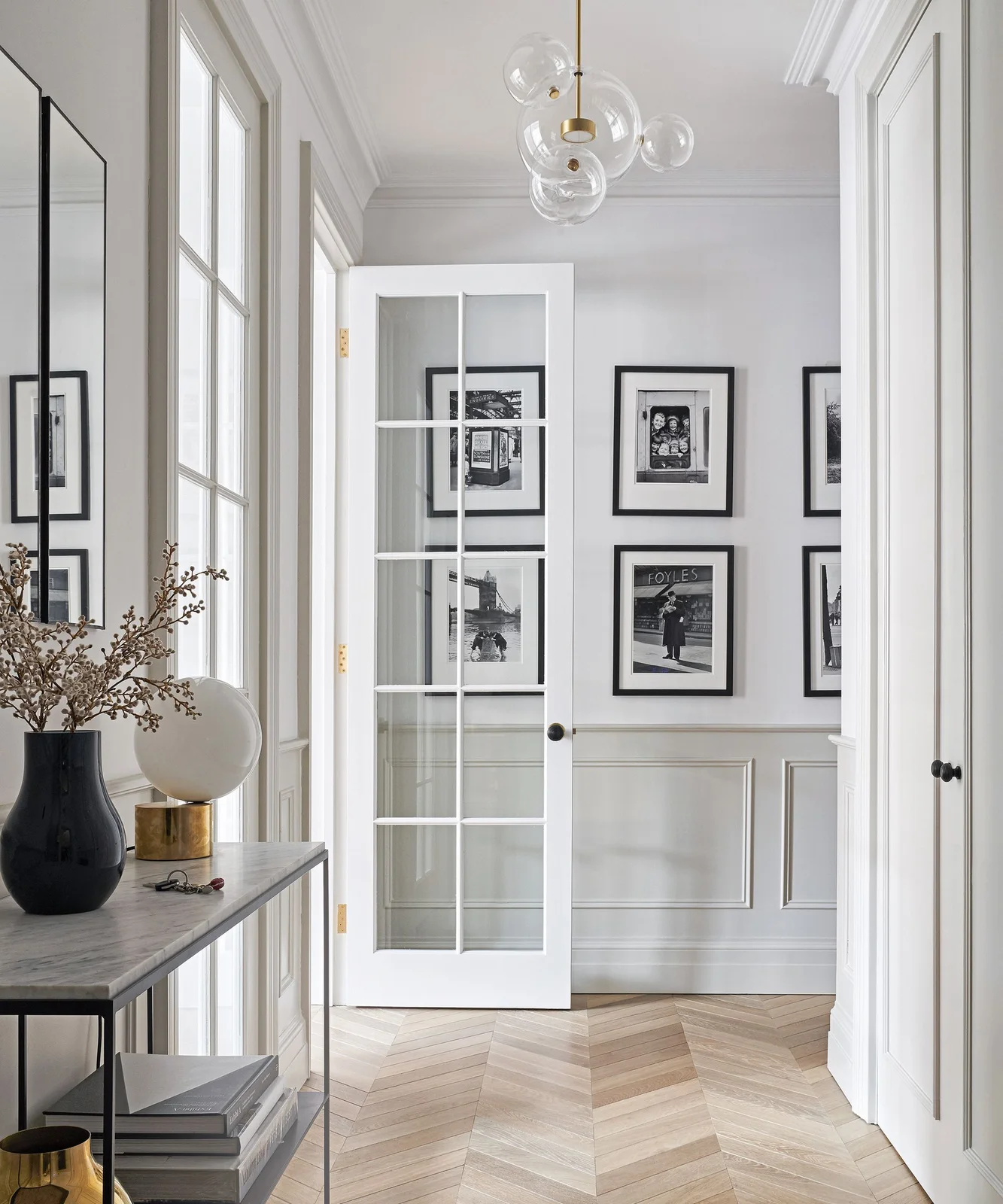 A pale hallway has white glazed doors, black-framed pictures and a slim console with a dark vase below clear globe lighting beside a herringbone floor
