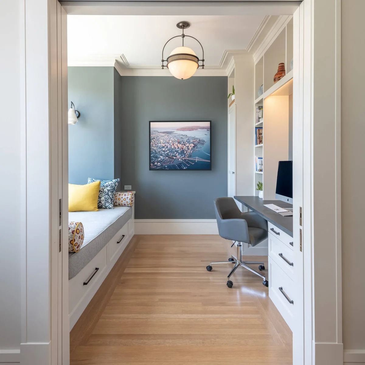 A gray-cushioned built-in daybed with lower drawers faces a desk in a blue-gray room with yellow accent cushions.