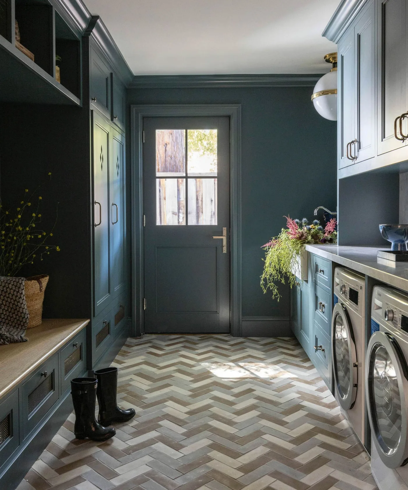 Dark blue cabinetry frames a glazed entry door, with a bench and boots on one side and laundry appliances beneath a counter on the other.