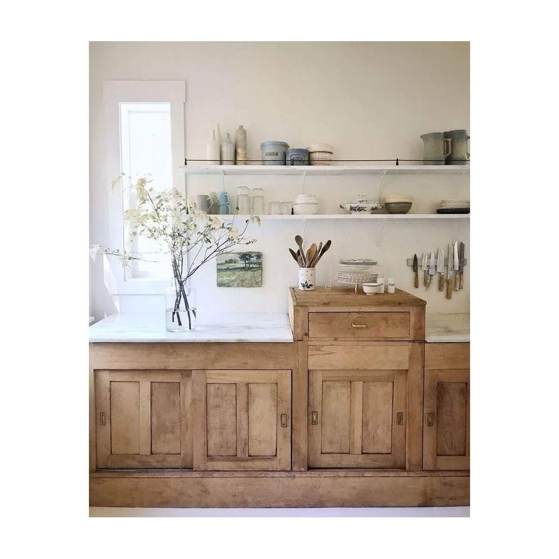 Plain white shelves holding crockery above worn wooden kitchen cupboards and pale counters