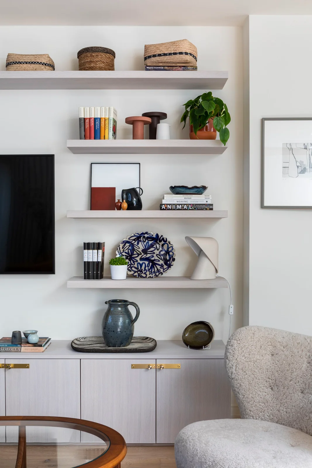 Pale floating shelves of different lengths hold books and ceramics above a matching cabinet with gold-colored handles beside a television.
