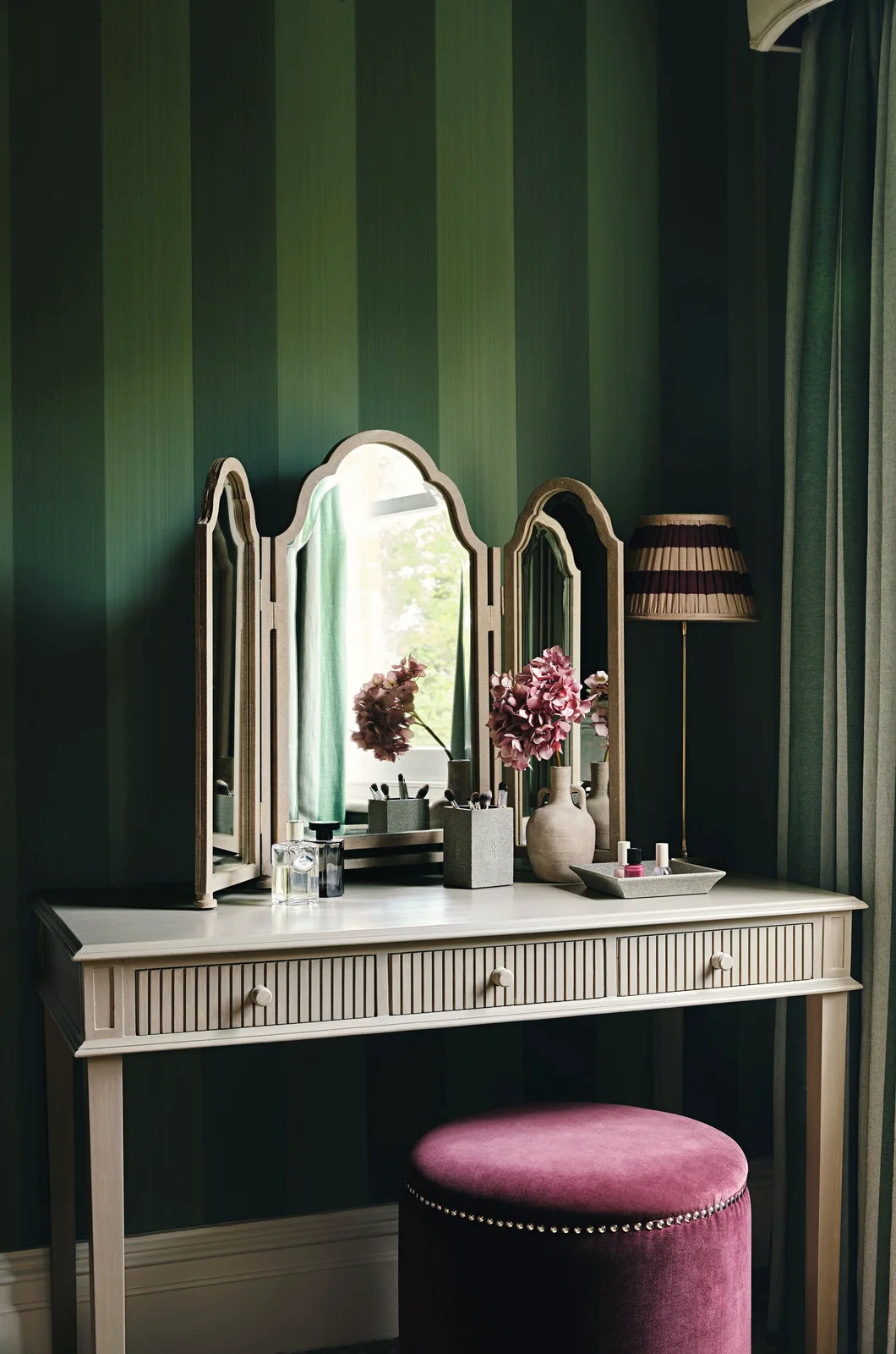 A pale dressing table and three-part mirror stand against green striped walls above a plum-colored stool