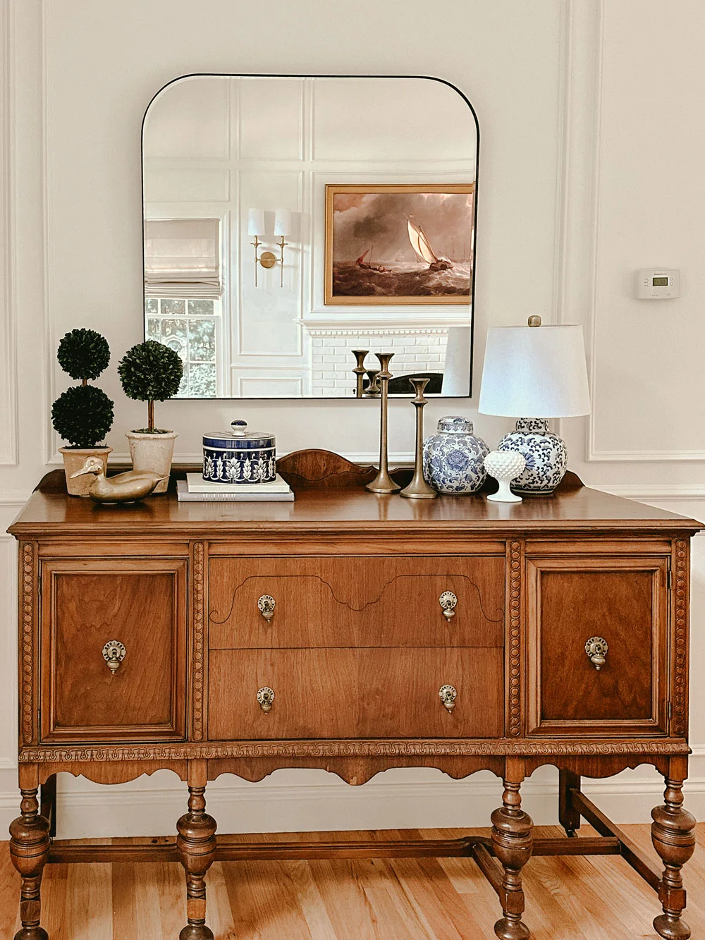 A carved timber sideboard stands beneath a large rounded rectangular mirror with a thin dark frame and blue-and-white vessels.