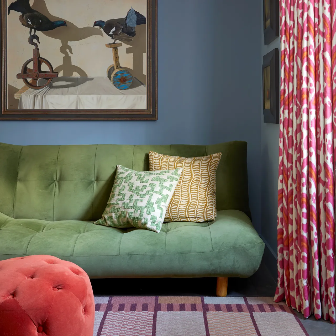 An olive green buttoned sofa sits beside a pink patterned curtain and red tufted footstool beneath framed artwork