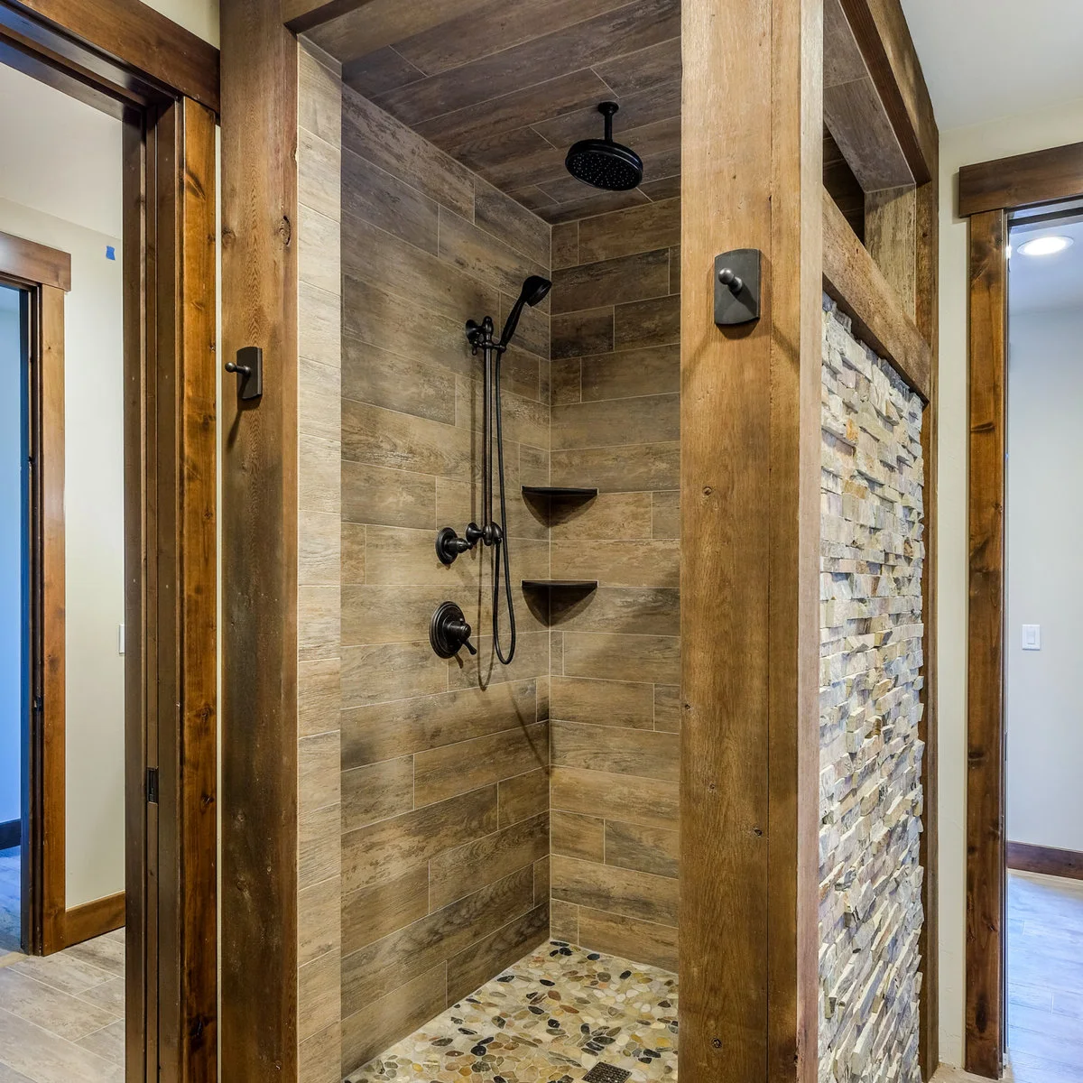A shower with wood-grain rectangular wall tiles, black fittings, two corner shelves and a pebble-patterned floor beside a timber-framed partition.