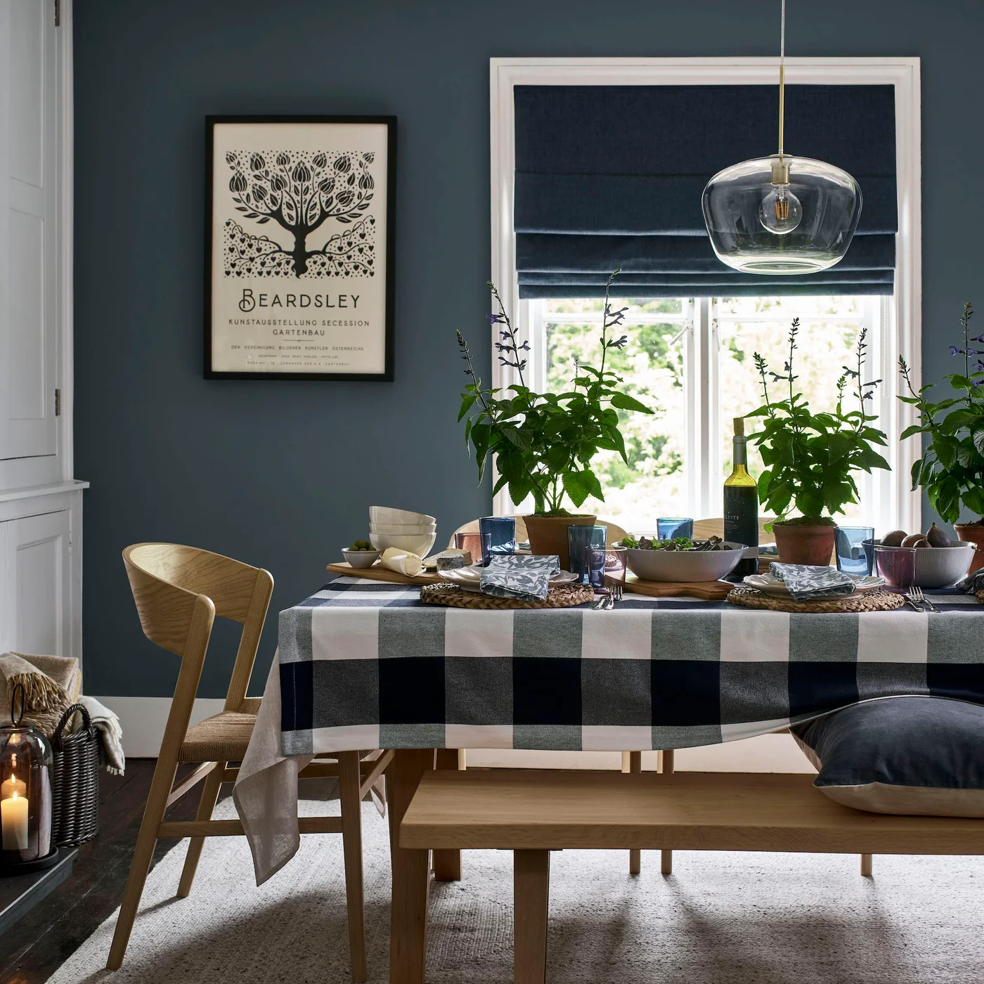 A navy Roman-style blind hangs above a dining table with a large checked cloth, timber bench and blue-gray walls.