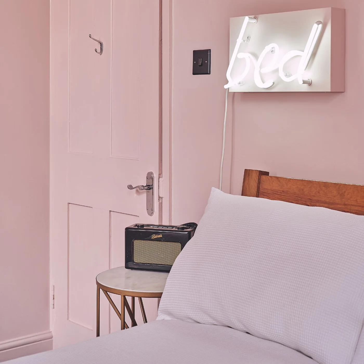 A pink bedroom wall and matching paneled door sit beside a wood bed, a small round bedside table, a radio and a white illuminated bed sign.