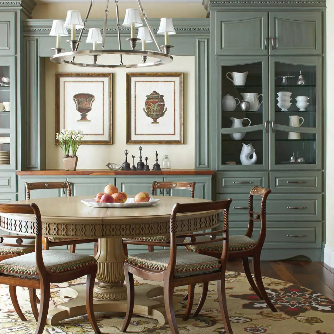 Floral patterned rug beneath a round pedestal dining table and carved wood chairs beside green cabinets
