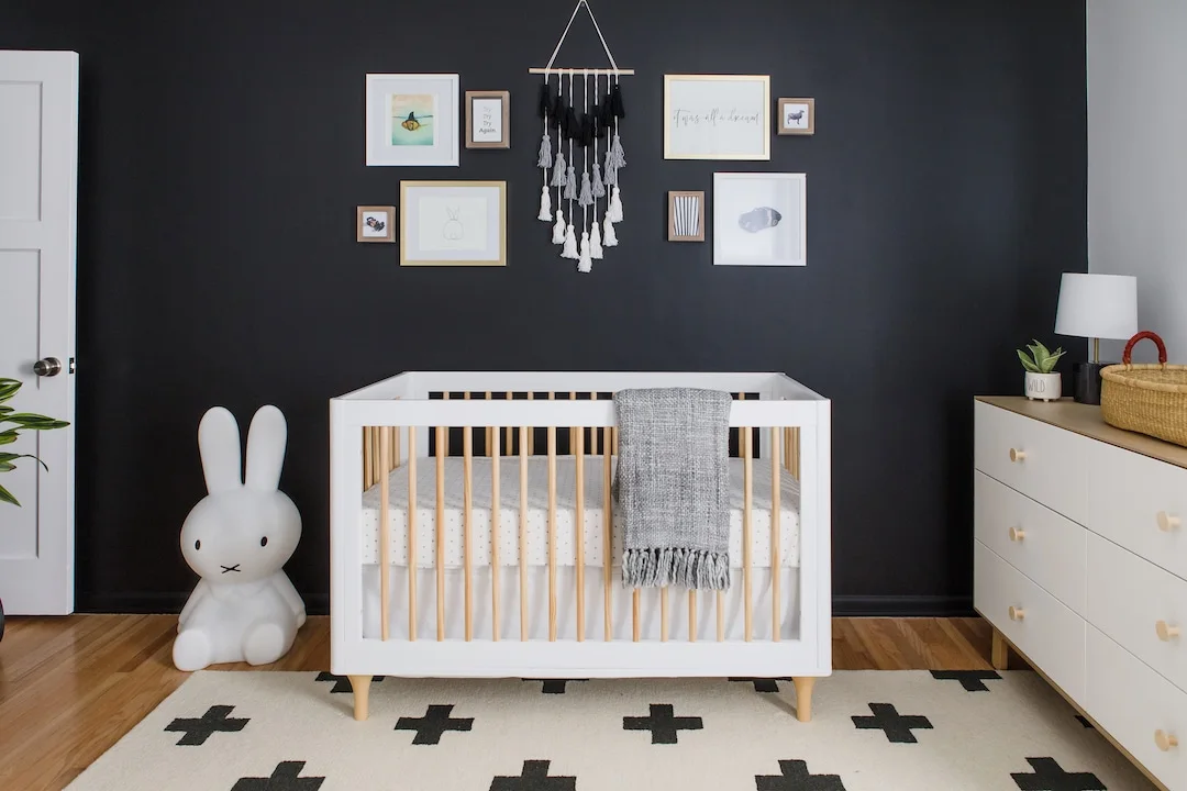 A pale nursery crib with warm slats stands below framed pictures and a tassel hanging on a dark wall beside pale drawers and a black-cross patterned rug