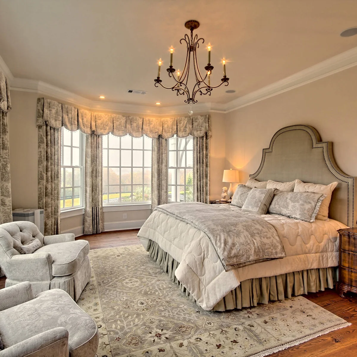 A pale traditional patterned rug runs beneath a bed and two light armchairs beside a bay window.