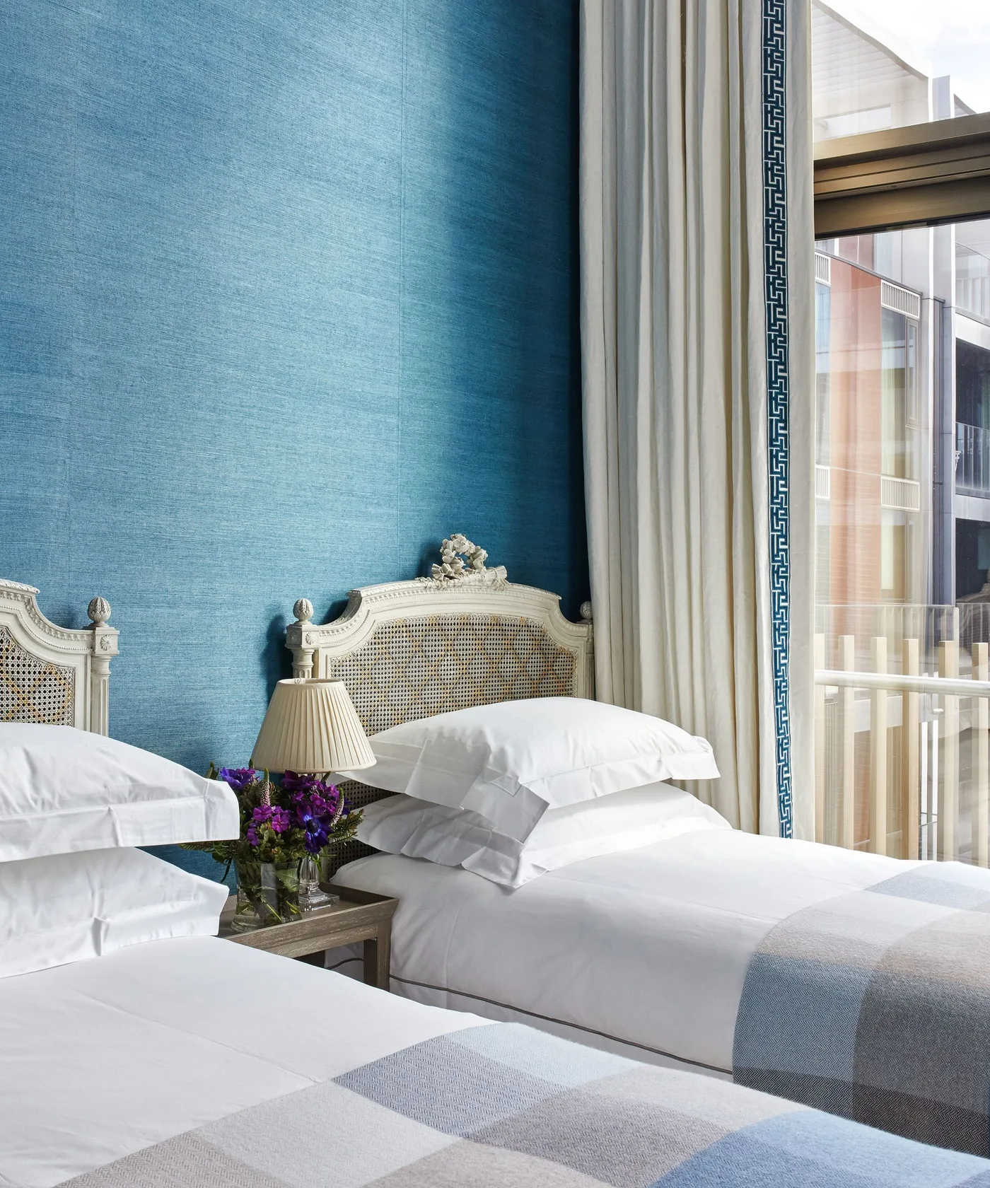 Two white woven headboards against a blue wall with white bedding, checked throws and edged curtains.