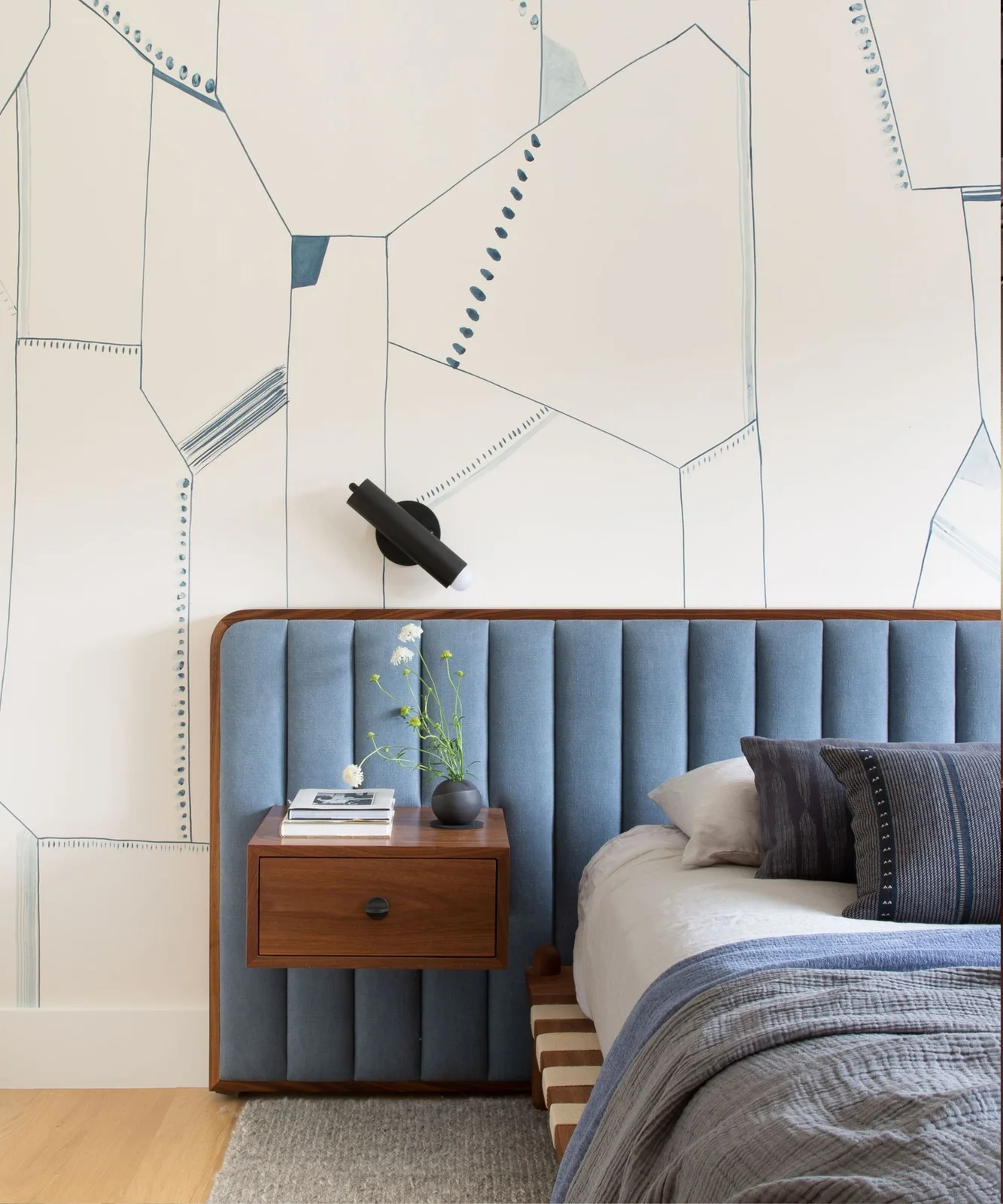 Large fine blue geometric lines cover a pale wall behind a blue channeled headboard, wood bedside drawer and black reading light.