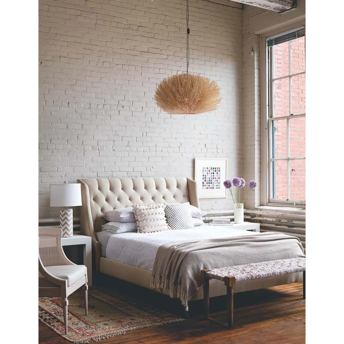 A pale tufted bed stands before a white brick wall beside a tall window, woven pendant and narrow upholstered bench