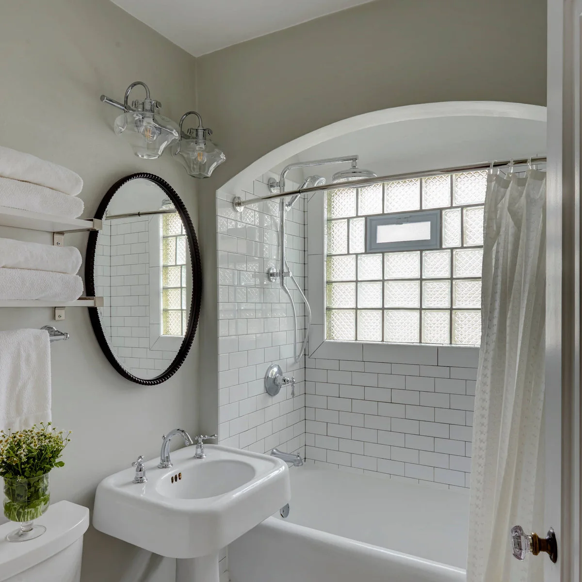 A white pedestal basin and dark oval mirror sit beside an arched bath opening with white tile and a glass-block window.