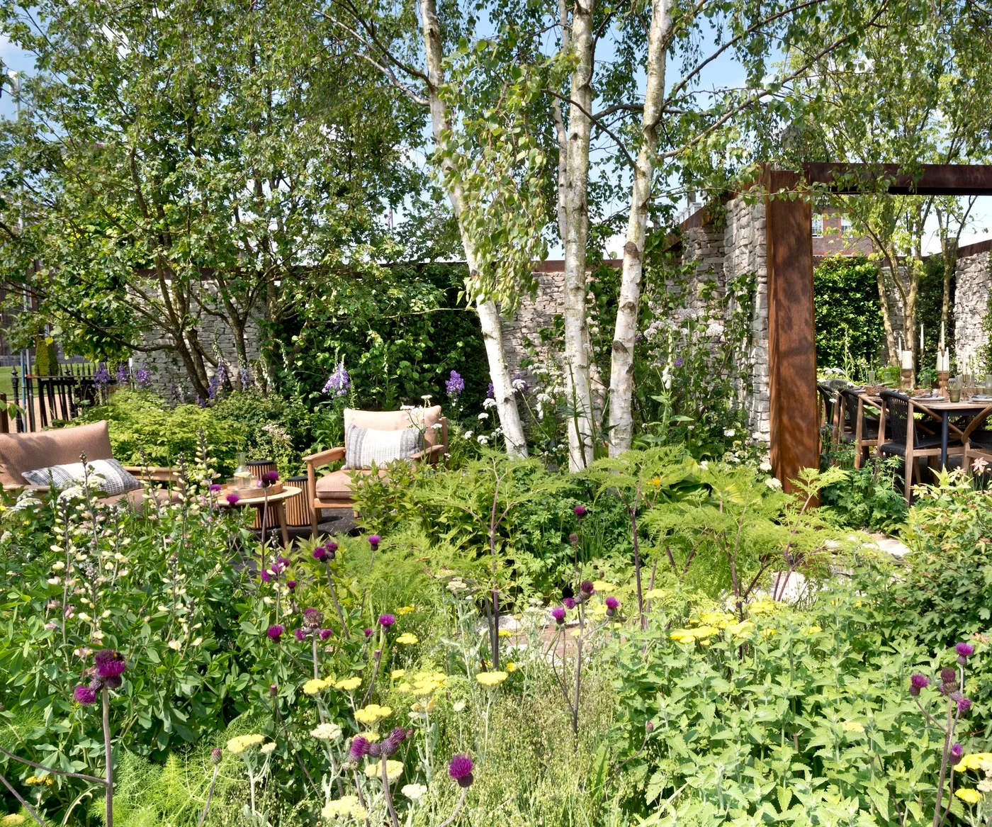 Two timber chairs sit beside a small table among layered planting, slender trees and stone walls