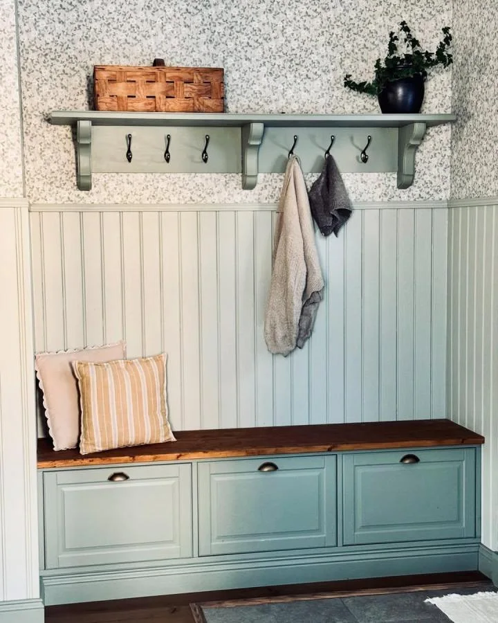 Pale green drawer-front storage with a dark wood top beneath a matching hook shelf and small patterned wallpaper.