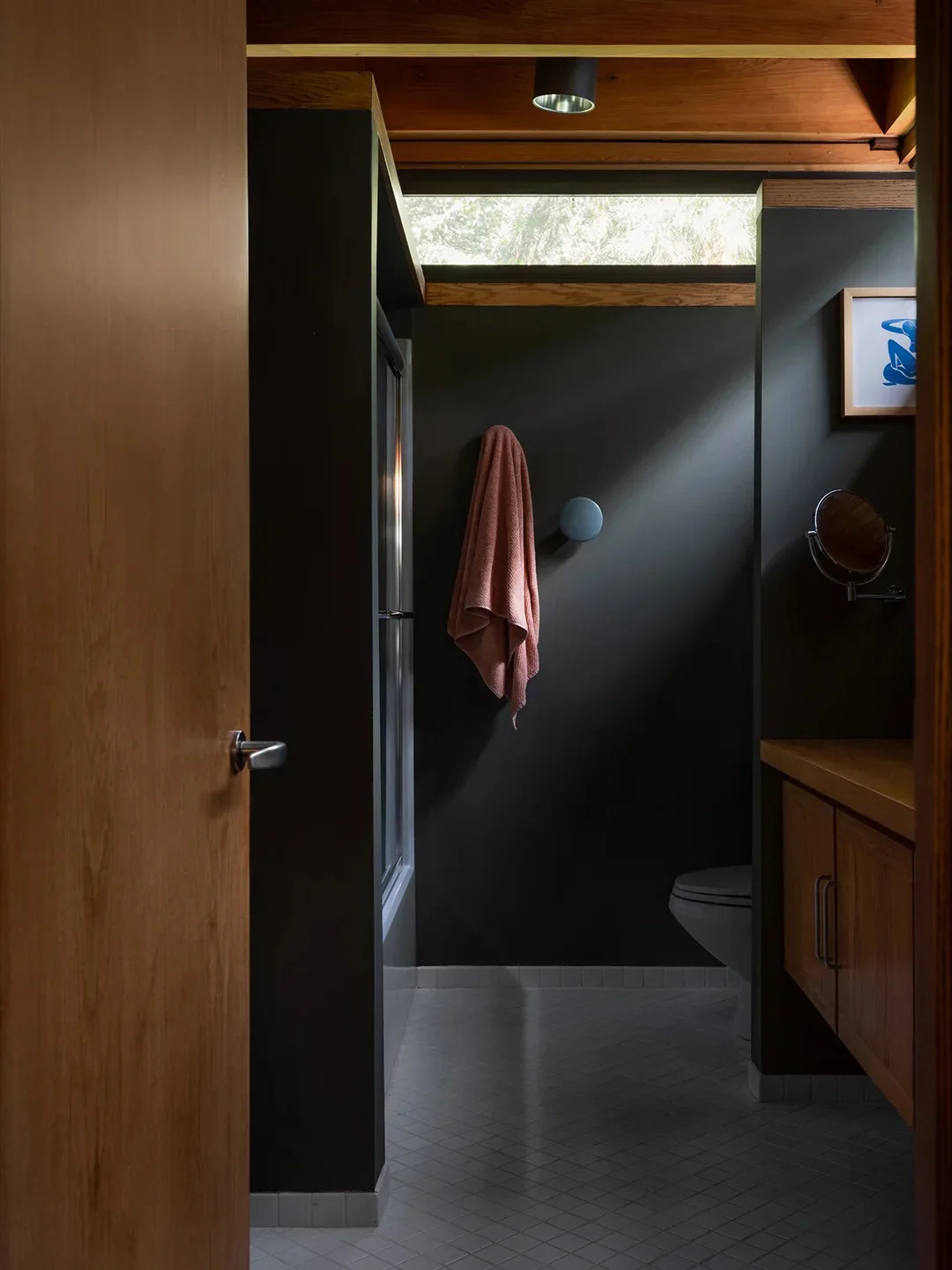 Dark bathroom walls sit beneath wood ceiling beams and high windows beside a wood door, gray patterned floor and dark shower frame