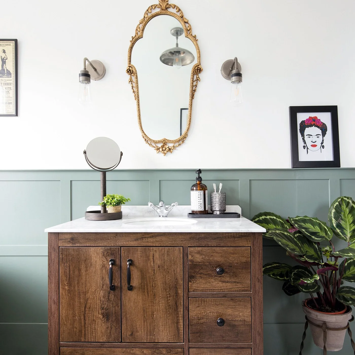 Sage lower-wall paneling sits behind a wood vanity, white countertop, ornate gold mirror and exposed-bulb wall lights.