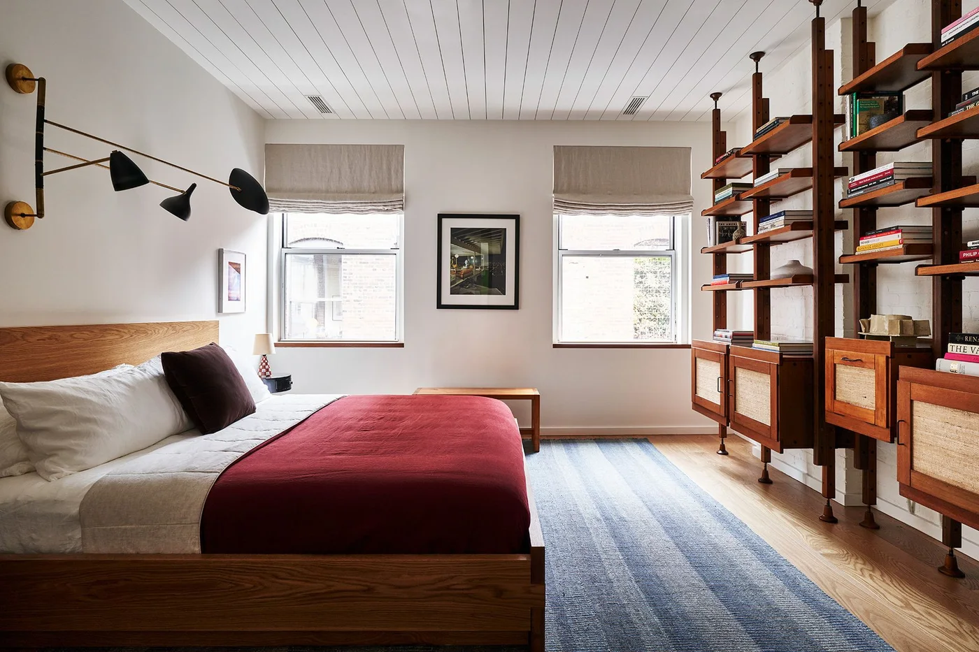 A bedroom has a simple wood bed with a red cover, tall wood-and-metal book shelving, two windows with pale shades and a long blue-gray rug.