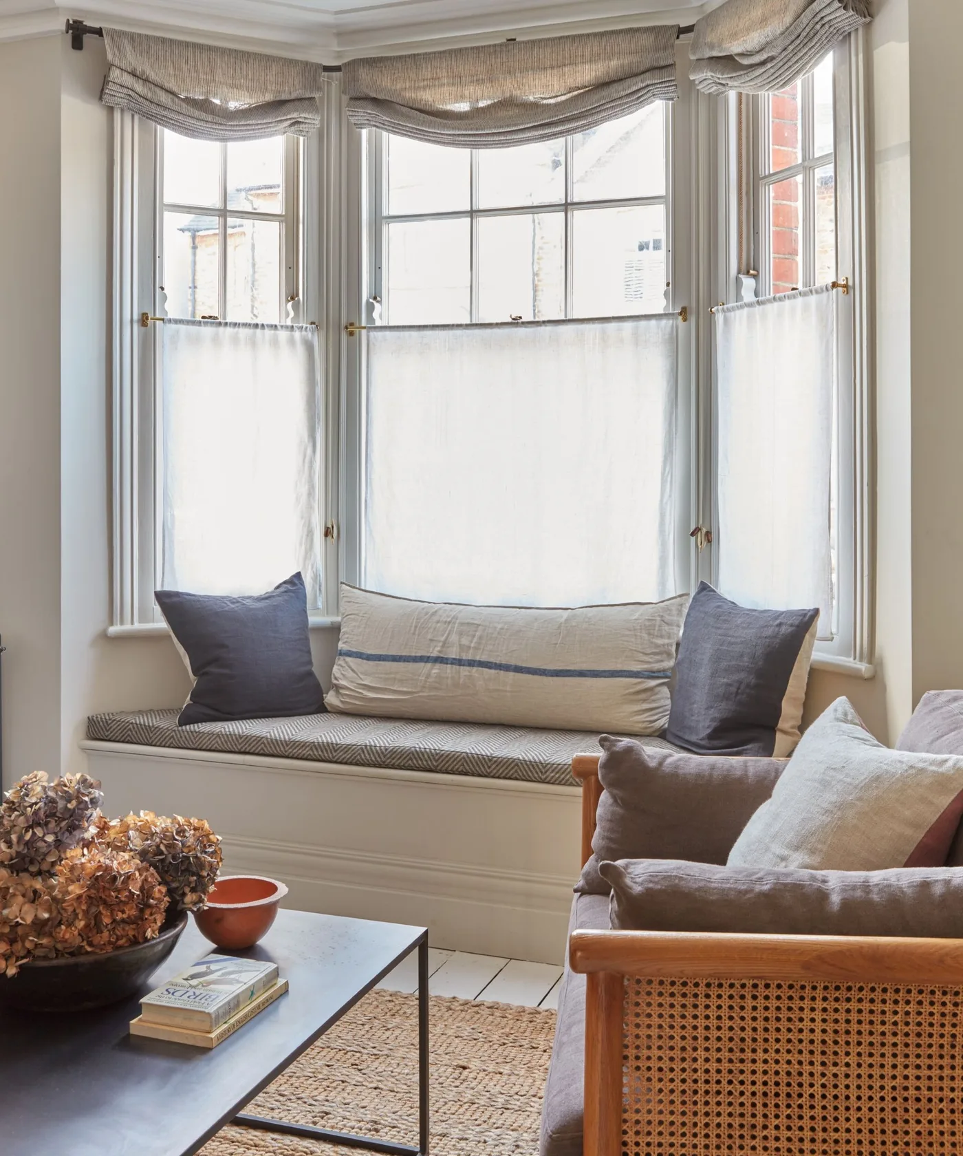 Pale bay-window bench with neutral cushions beside a woven-backed wood chair and simple dark coffee table