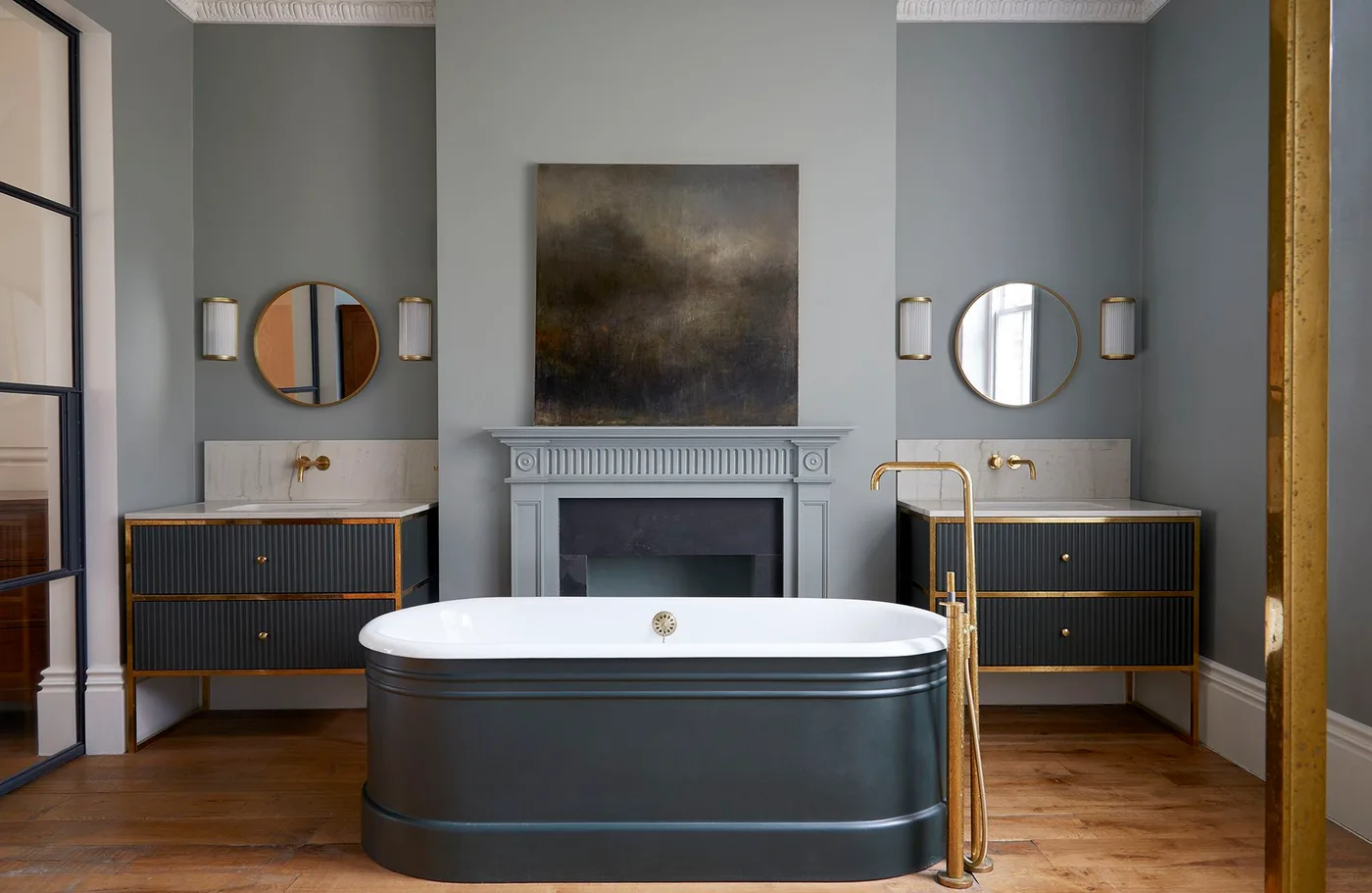Charcoal freestanding tub between two dark ribbed vanities with warm metal trim, gray walls, round mirrors, fireplace surround, and wooden floor.