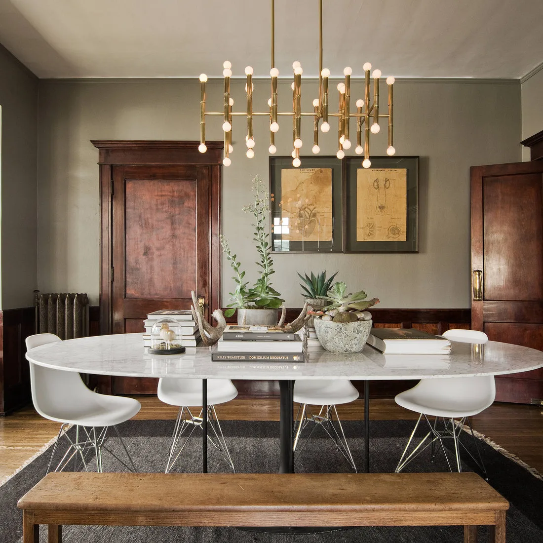 Pale oval dining table with white chairs and a wood bench beneath a gold-toned branching light
