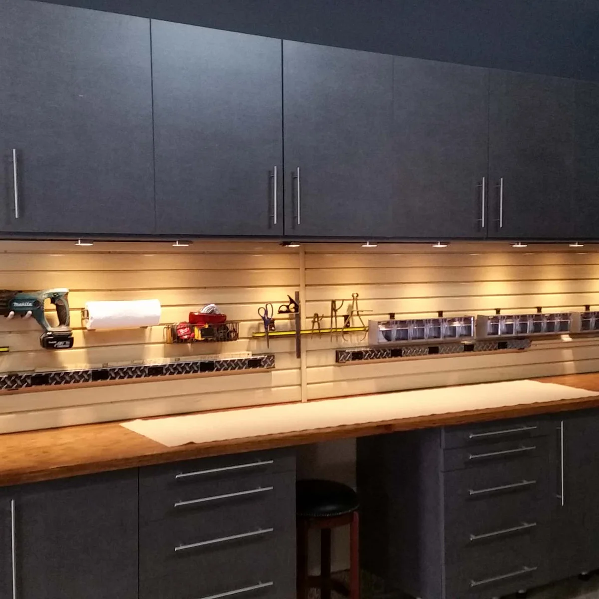 Long wooden garage worktop with dark cabinets above and below, illuminated tools and small-parts containers on a slat wall.