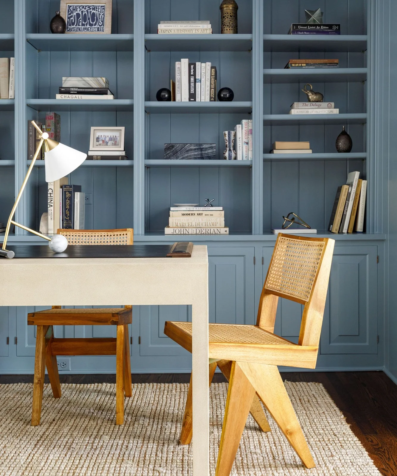 Blue open shelves and lower cupboards form a backdrop to a pale desk with wood chairs, a light task lamp and a woven rug.