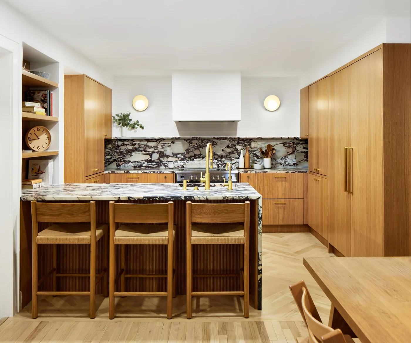 Plain wooden cabinets surround a black-and-white veined backsplash and island counter beside three wooden stools and small round wall lights