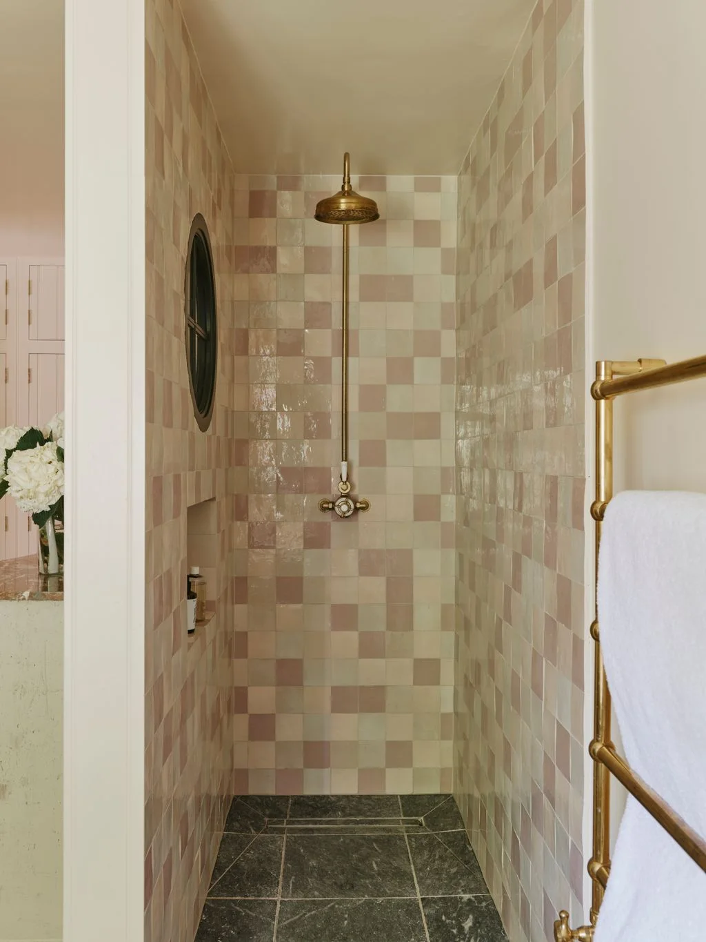 A narrow open shower alcove has pink and cream checker tiles, a round dark window and a brass-colored column shower.