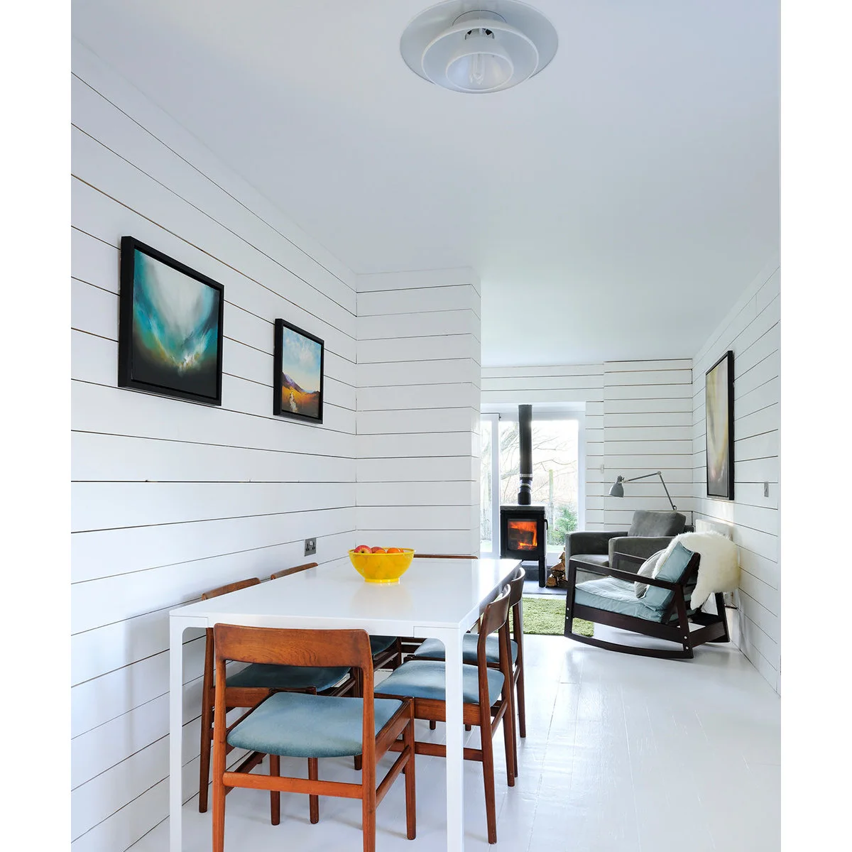 Very narrow white paneled room with a white dining table, wooden chairs and two armchairs beside a stove at the far end.