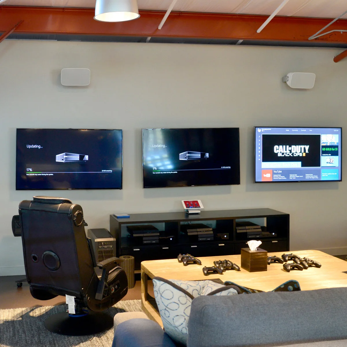 Three wall-mounted gaming screens above a black console, with a black gaming chair, a low controller table and a gray sofa.