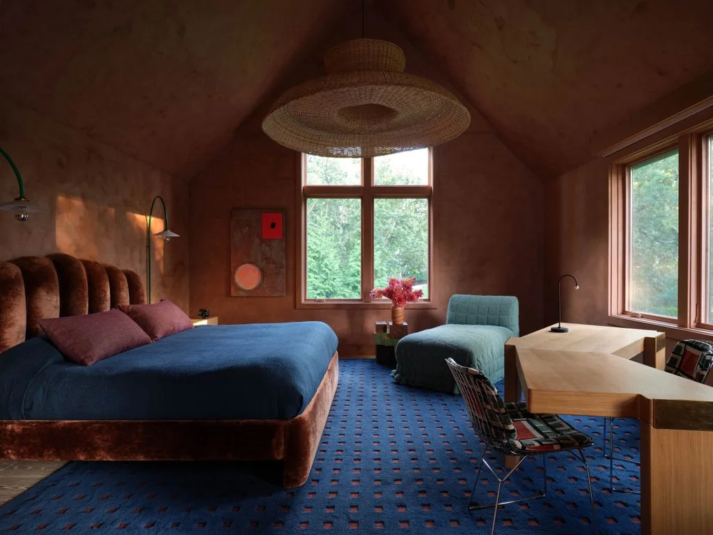 A blue-covered bed and pale blue chair stand within warm reddish walls beside an angular wood desk, broad windows and a large woven pendant