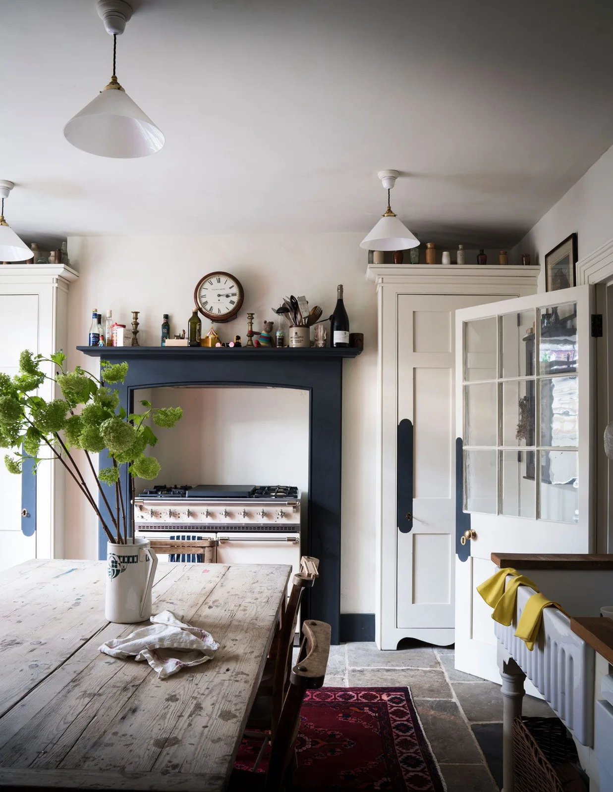 A worn plank dining table stands before a cream range cooker with a navy surround and white kitchen cabinets.