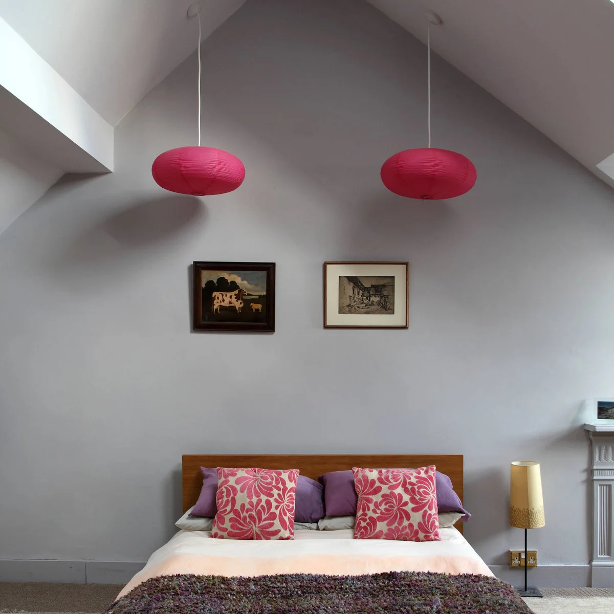 Gray bedroom with two bright pink hanging shades and pink floral cushions on a wood-colored bed