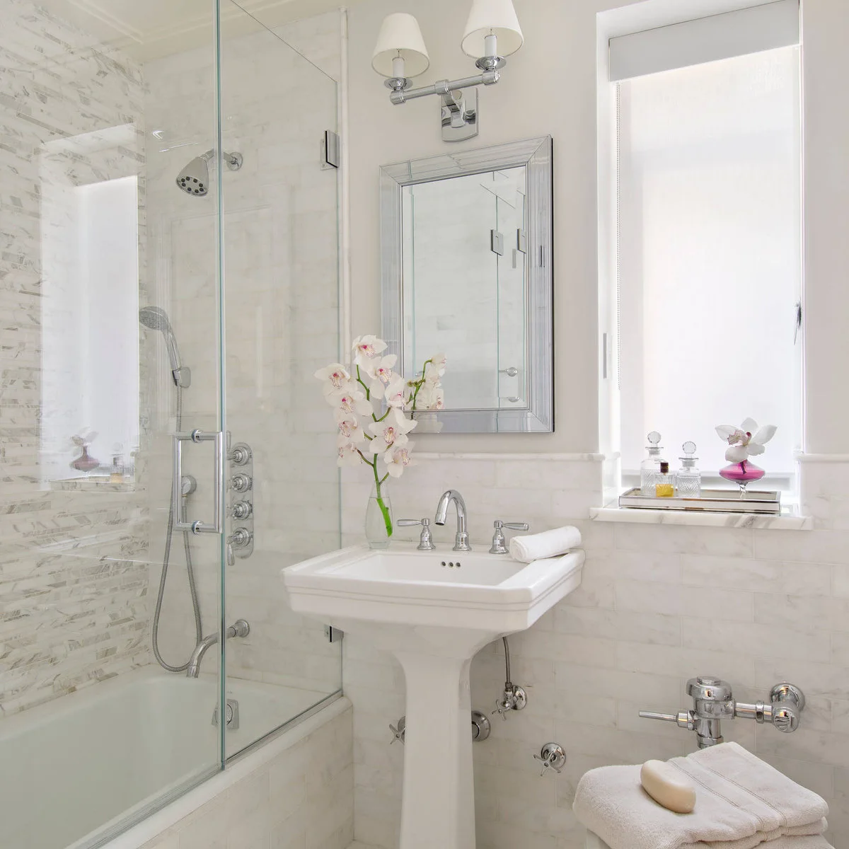 A white pedestal sink stands between a glass shower enclosure and a bright window in a pale tiled bathroom.