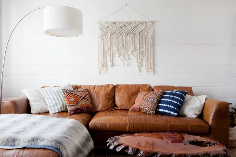 Brown leather sectional with patterned cushions, pale throw, macrame wall hanging and curved floor lamp.