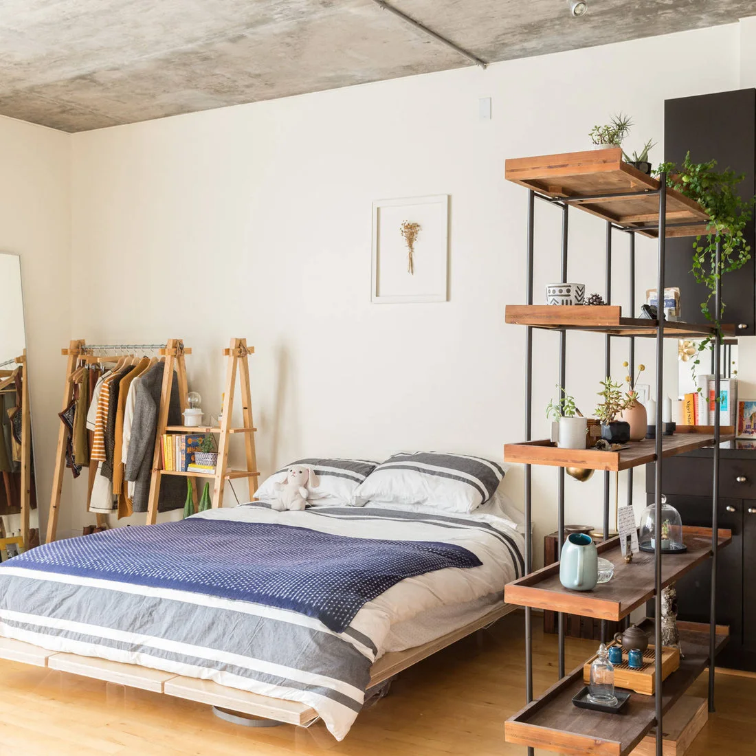 A low wooden bed with striped pale bedding and a blue throw sits beside open wood-and-black-frame shelves beneath a rough ceiling