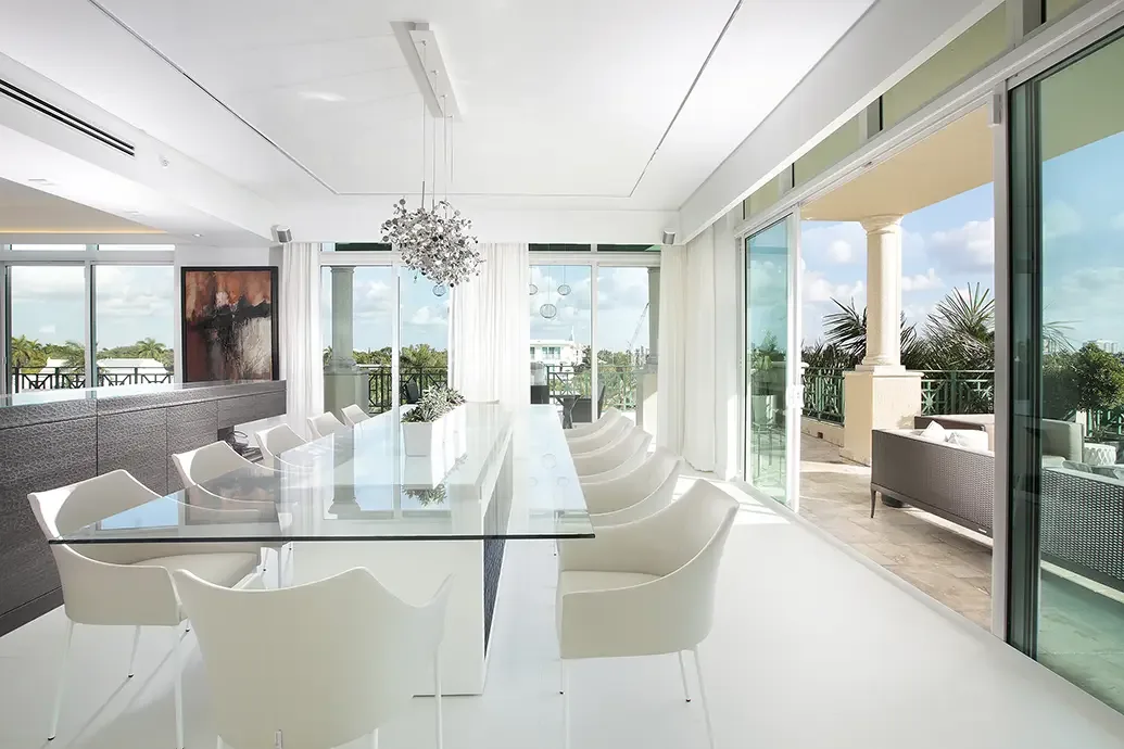 Long clear glass dining table on a pale central base surrounded by white chairs beside sliding doors