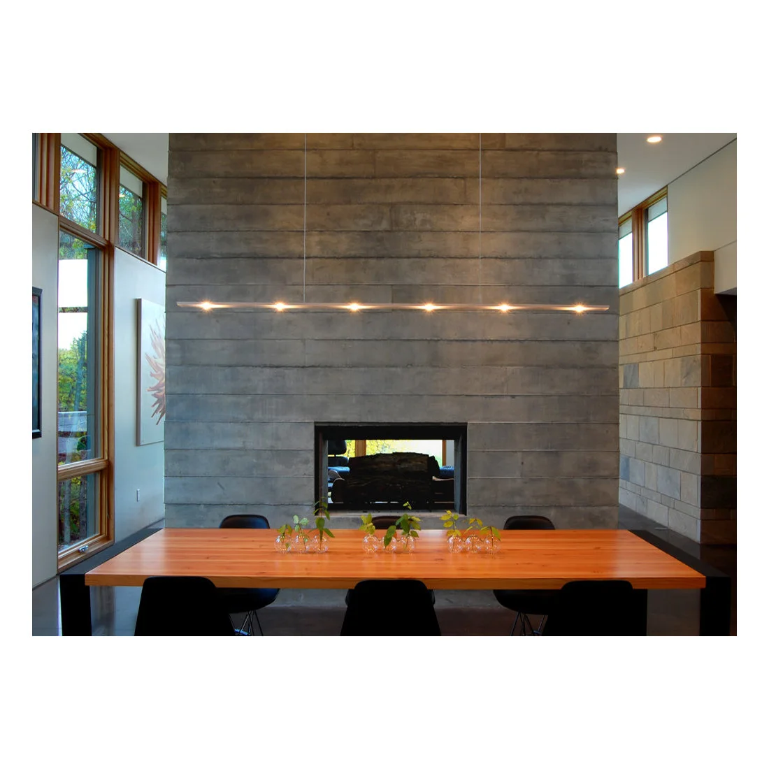 A warm wood dining table and dark chairs sit beneath a thin linear pendant beside a broad gray horizontally lined fireplace wall and small plant vases