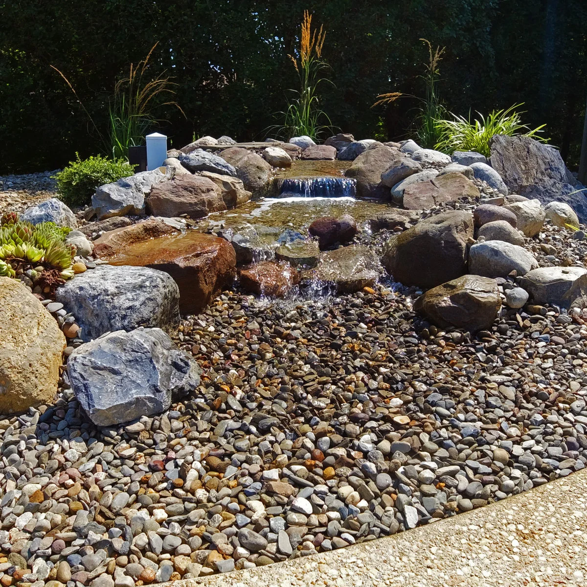 A low garden cascade between large rocks, with a broad pebble-covered lower area and sparse grasses behind.