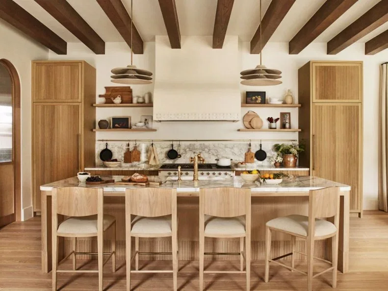 Tall fluted wood cabinets flank a pale hood and open shelves behind a wood island with four matching stools.