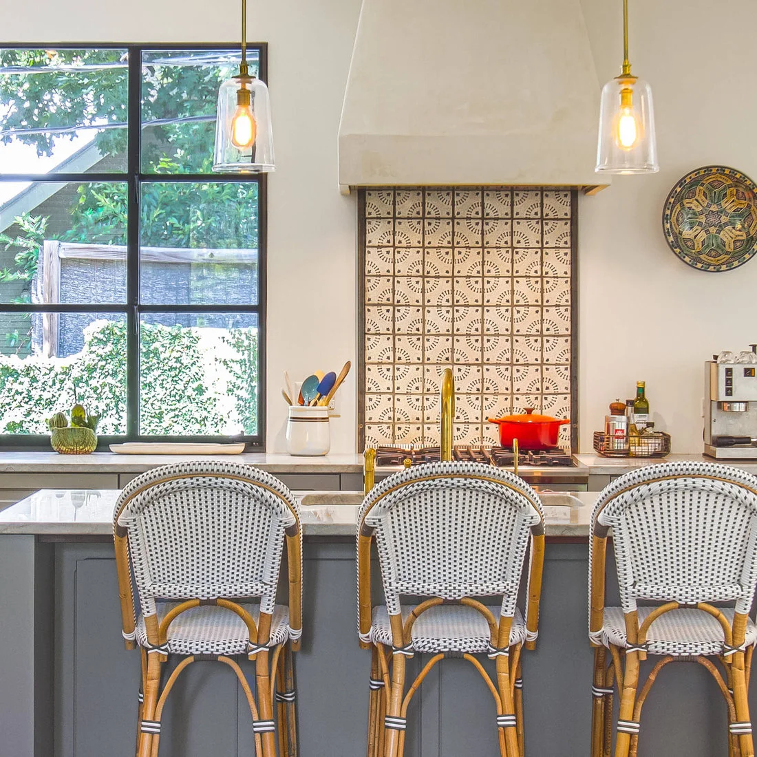 A cream-and-dark patterned range backsplash sits beneath a pale hood behind woven counter stools and gray cabinets.